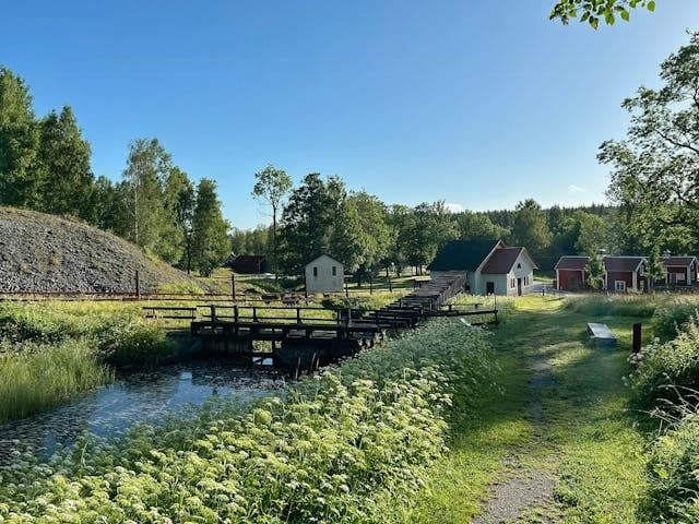 Vacker utsikt över Villa Pershyttans lantliga omgivningar med bäck, träbro och traditionella röda stugor omgivna av grönska och träd.